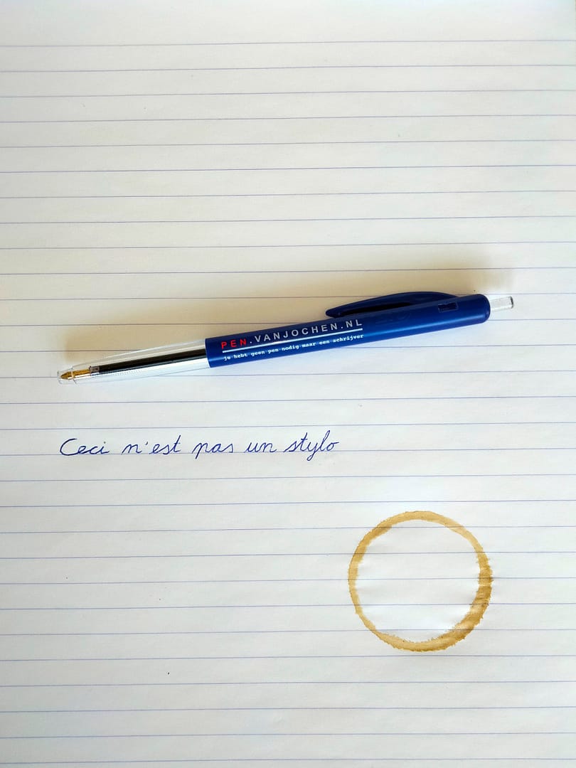 een balpen met opschrift 'Pen.vanjochen.nl Je hebt geen pen nodig maar een schrijver' ligt op een kladblok. Daaronder staat in een schools handschrift 'Ceci n'est pas un stylo'. Op het papier zit een koffievlek